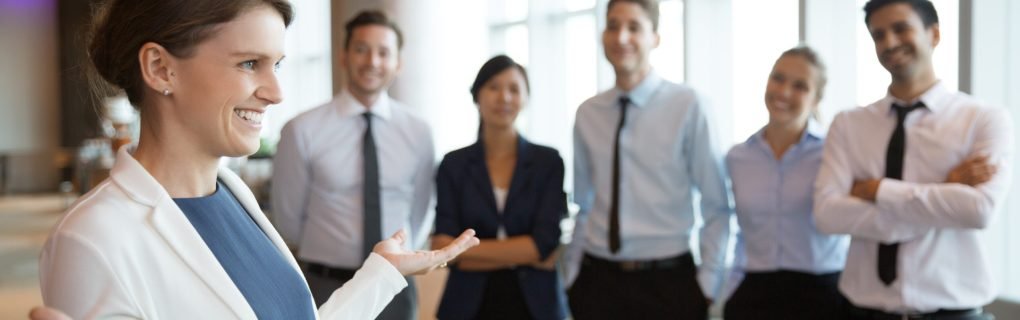 Portrait of smiling businesswoman with open hands gesture standing in office hall, her business team on background